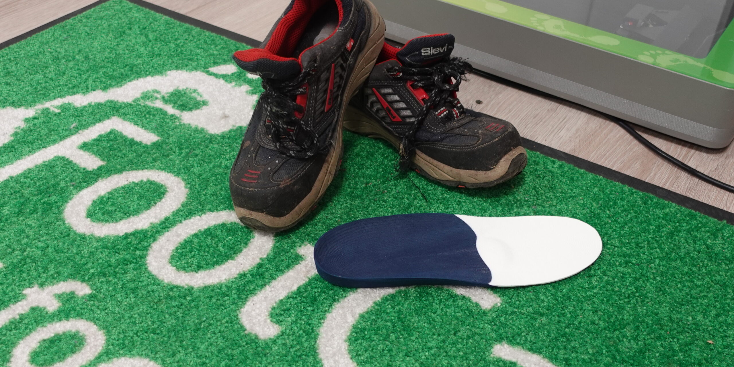 FootCare-tukipohjallinen ja turvajalkineet Reijolan Fysioterapiassa Joensuussa FootCare-tukipohjallinen asetettuna turvajalkineiden viereen FootCare-logolla varustetulla alustalla Reijolan Fysioterapiassa Joensuussa