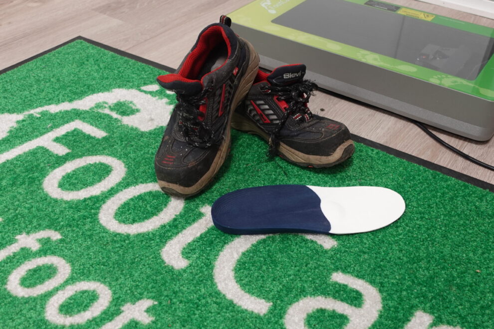 FootCare-tukipohjallinen asetettuna turvajalkineiden viereen FootCare-logolla varustetulla alustalla Reijolan Fysioterapiassa Joensuussa