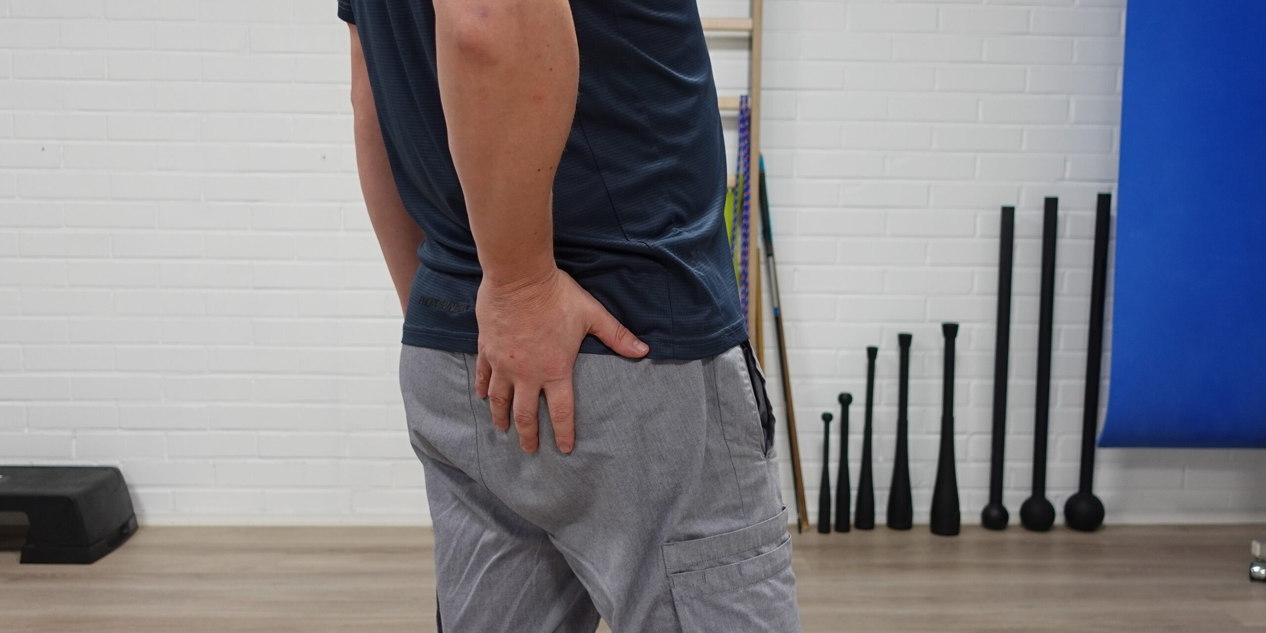 Fysioterapeutti seisoo takaviistosta kuvattuna ja pitää kättä alaselän ja pakaran päällä samalla kun keventää jalkaa. Kuva havainnollistaa iskiasoireita ja hermopainetta, joka voi heijastua jalkaan.